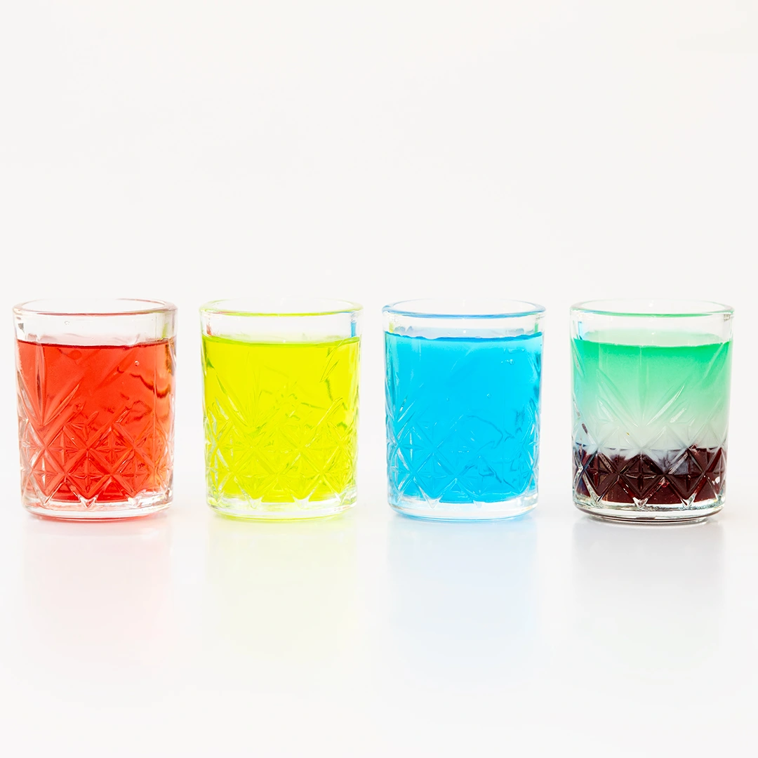 Si vedono quattro bicchieri di shots pieni. Ogni bicchiere ha un colore diverso: rosso, giallo, azzurro e verde/bianco